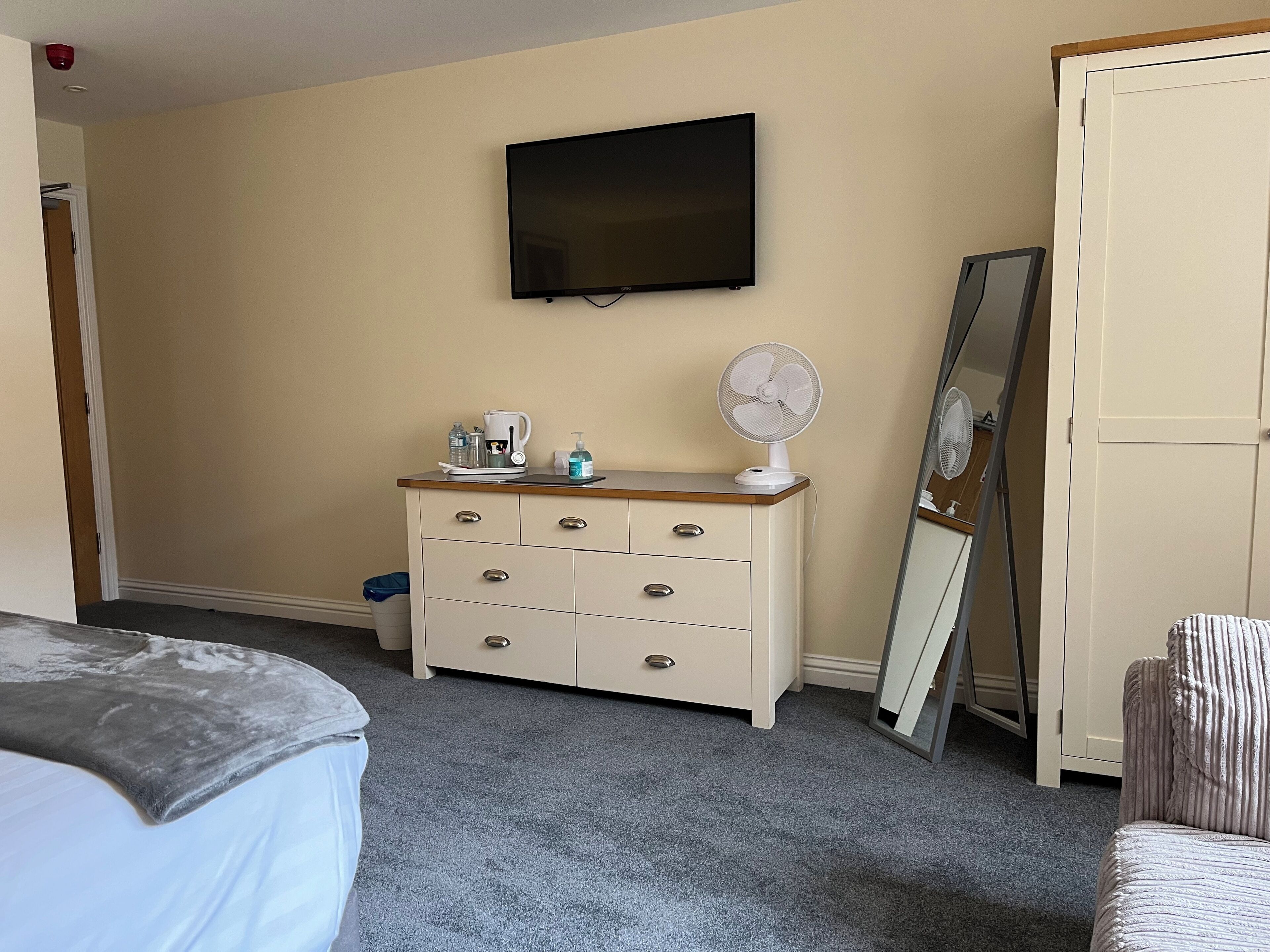 A bedroom with a bed, a dresser with a mirror, a television on the wall, and a small fan on the dresser.