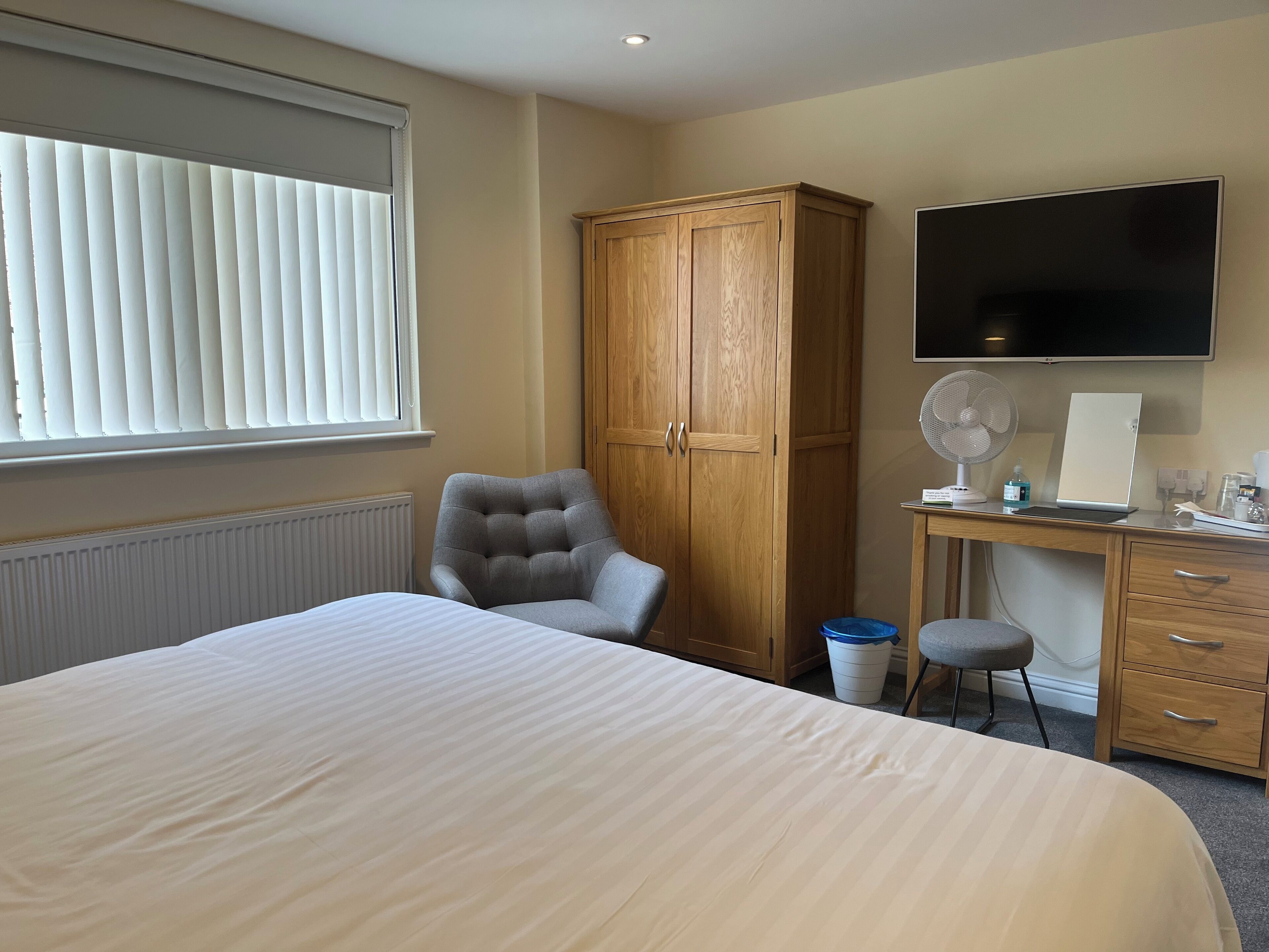 Une chambre d’hôtel avec un grand lit, une armoire en bois, un bureau avec un ordinateur, une chaise, une télévision et une fenêtre avec des stores.