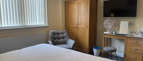 Une chambre d’hôtel avec un grand lit, une armoire en bois, un bureau avec un ordinateur, une chaise, une télévision et une fenêtre avec des stores.