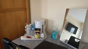 Una habitación de hotel con una puerta de madera, una mesa con una bandeja que contiene una cafetera, botellas de agua, un dispensador de gel antibacterial, una taza amarilla pequeña y una caja de pañuelos.
