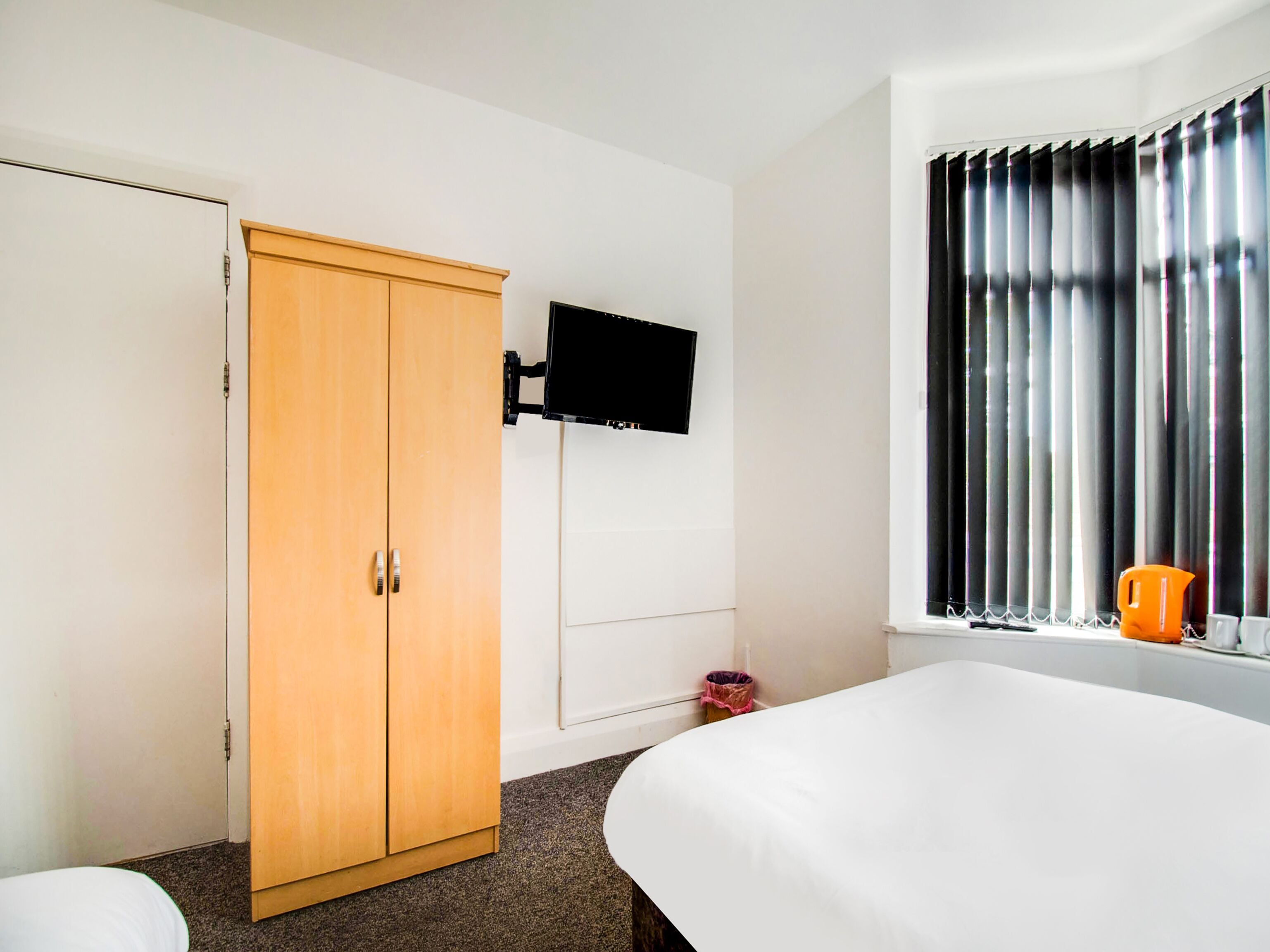A hotel room with a wooden wardrobe, a flat-screen TV mounted on the wall, a window with blinds, and a bed with a white comforter.