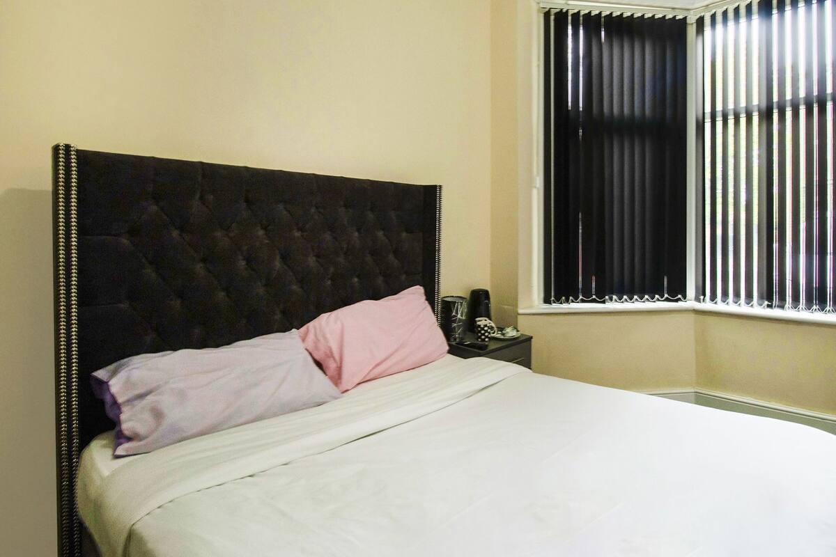 A bed with a tufted headboard, a bedside table with a phone and a cup, and a window with vertical blinds.