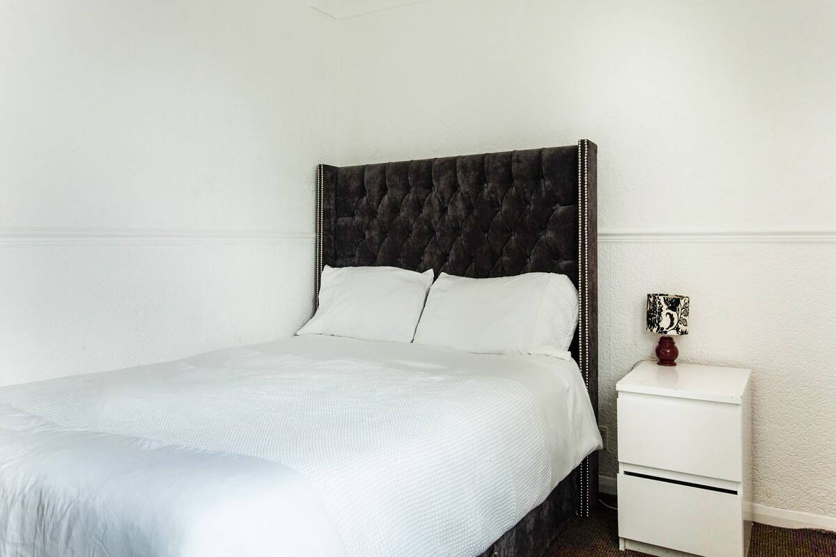 A neatly made bed with a dark headboard, a white bedside table with a decorative item, and a white wall.