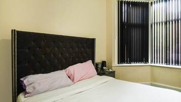A bedroom with a tufted headboard, a bed with white bedding and pink pillows, a bedside table with a phone and a cup, and a window with vertical blinds.