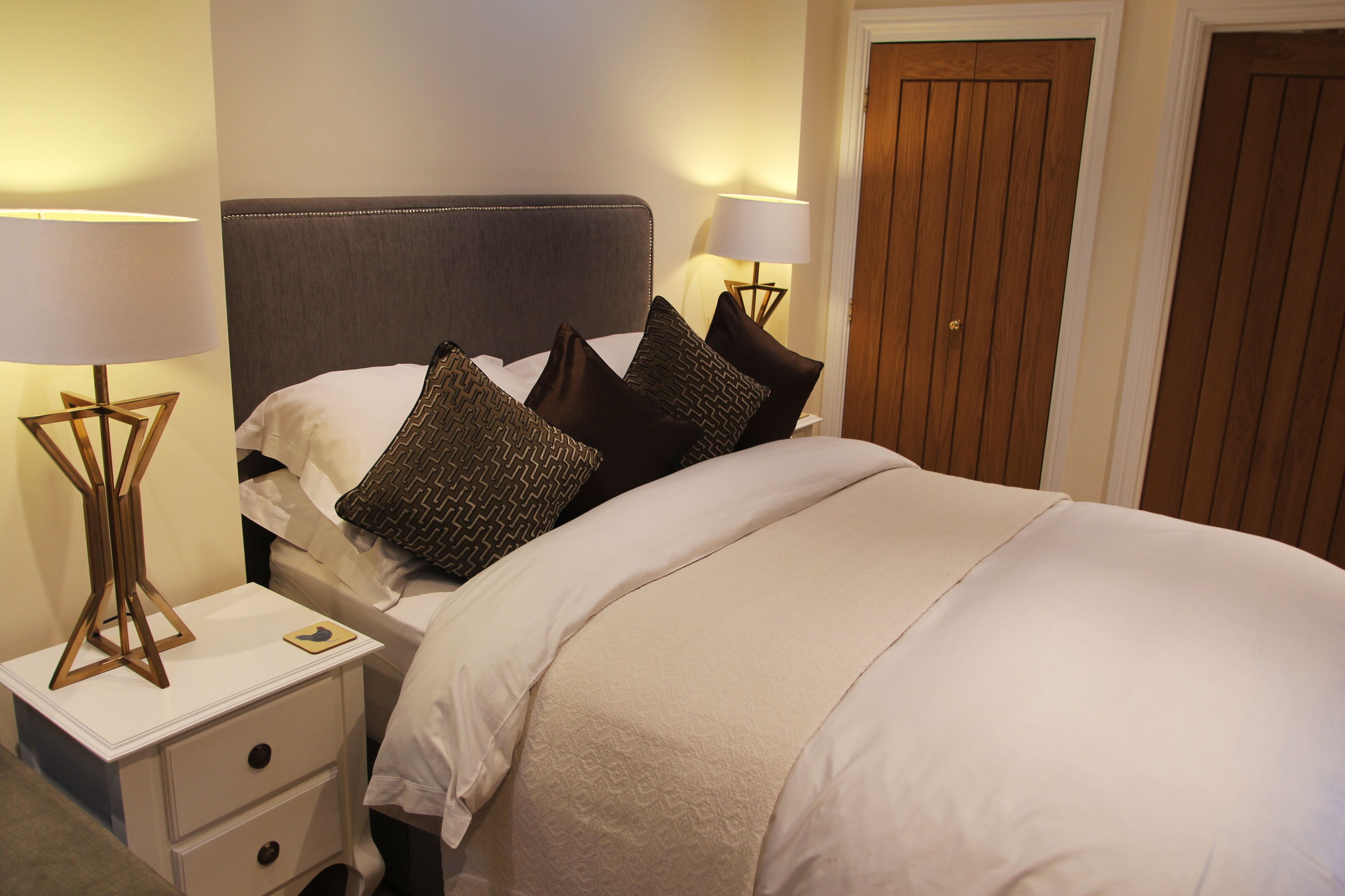 A neatly made bed with a grey headboard, two bedside lamps, and a nightstand with drawers.