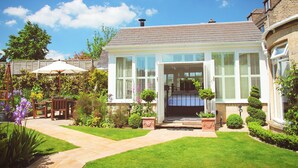 A two-story house with a well-maintained garden, a patio with outdoor furniture, and a clear blue sky. - Croft House Guest Suite (Stroud)