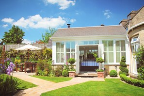 A two-story house with a well-maintained garden, a patio with outdoor furniture, and a clear blue sky. - Croft House Guest Suite (Stroud)