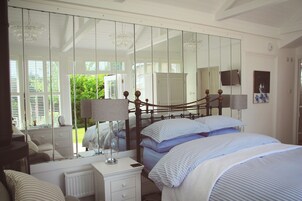 Un dormitorio con un espejo grande, una cama con ropa de cama azul, una mesita de noche con una lámpara y vistas al exterior a través de la ventana.