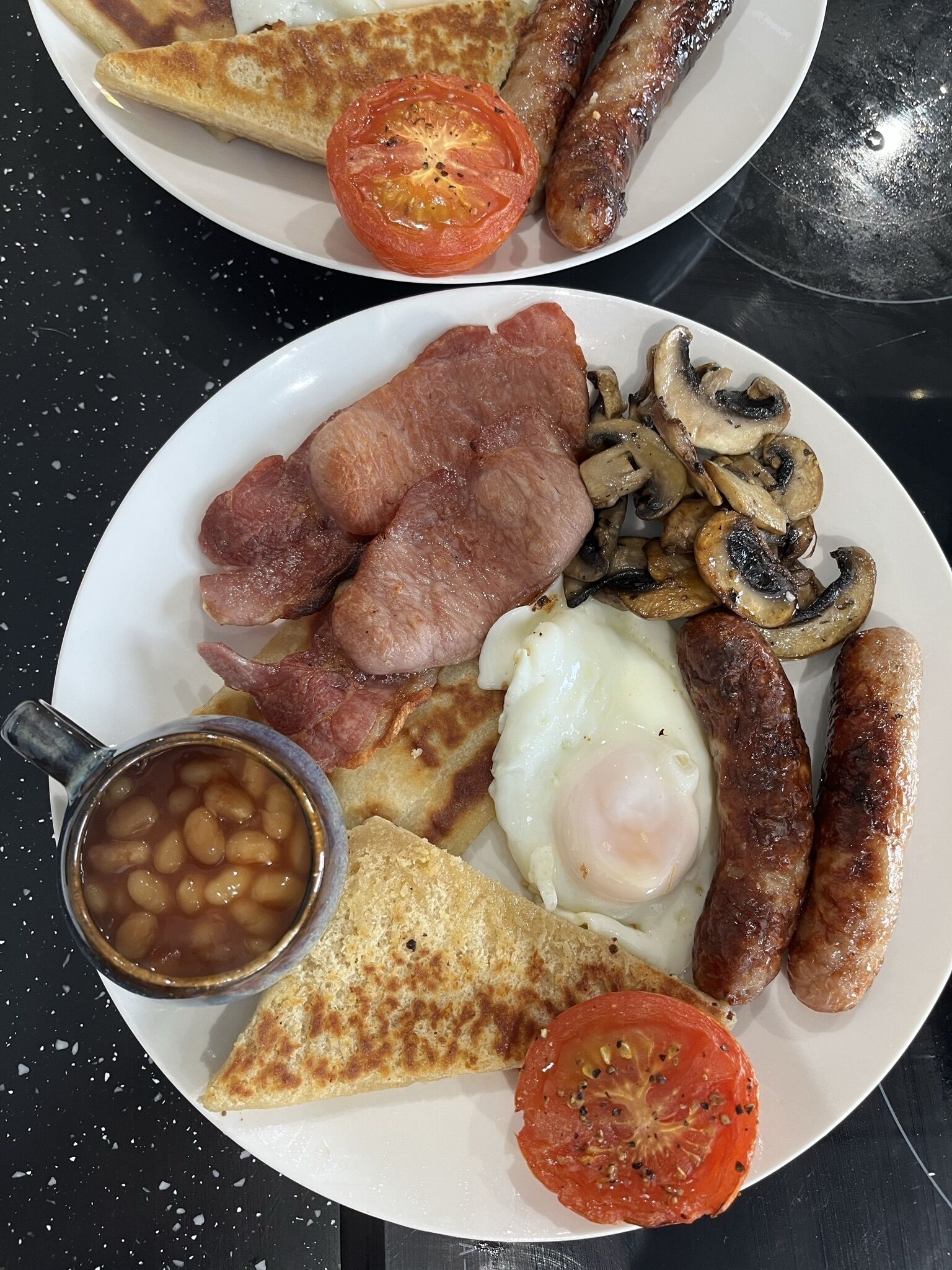 Un plato de desayuno con huevos, salchichas, tocino, champiñones, frijoles, tostadas y tomate.