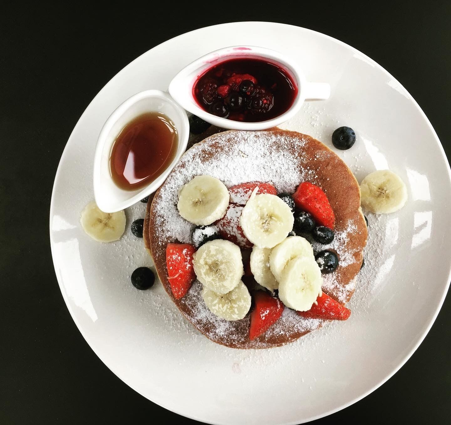 Ein Teller mit Pfannkuchen, belegt mit Bananenscheiben, Erdbeeren und Blaubeeren, dazu zwei kleine Schalen Ahornsirup und Puderzucker.