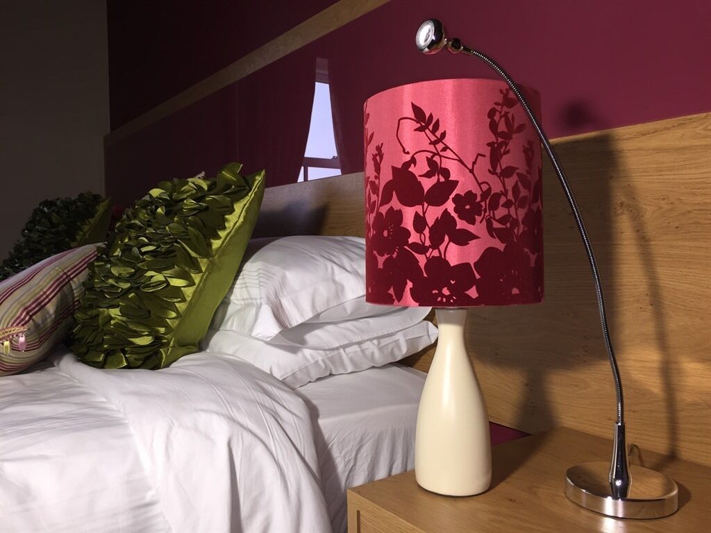 Una cama con ropa blanca, una almohada decorativa verde y una lámpara floral roja sobre una mesita de noche.