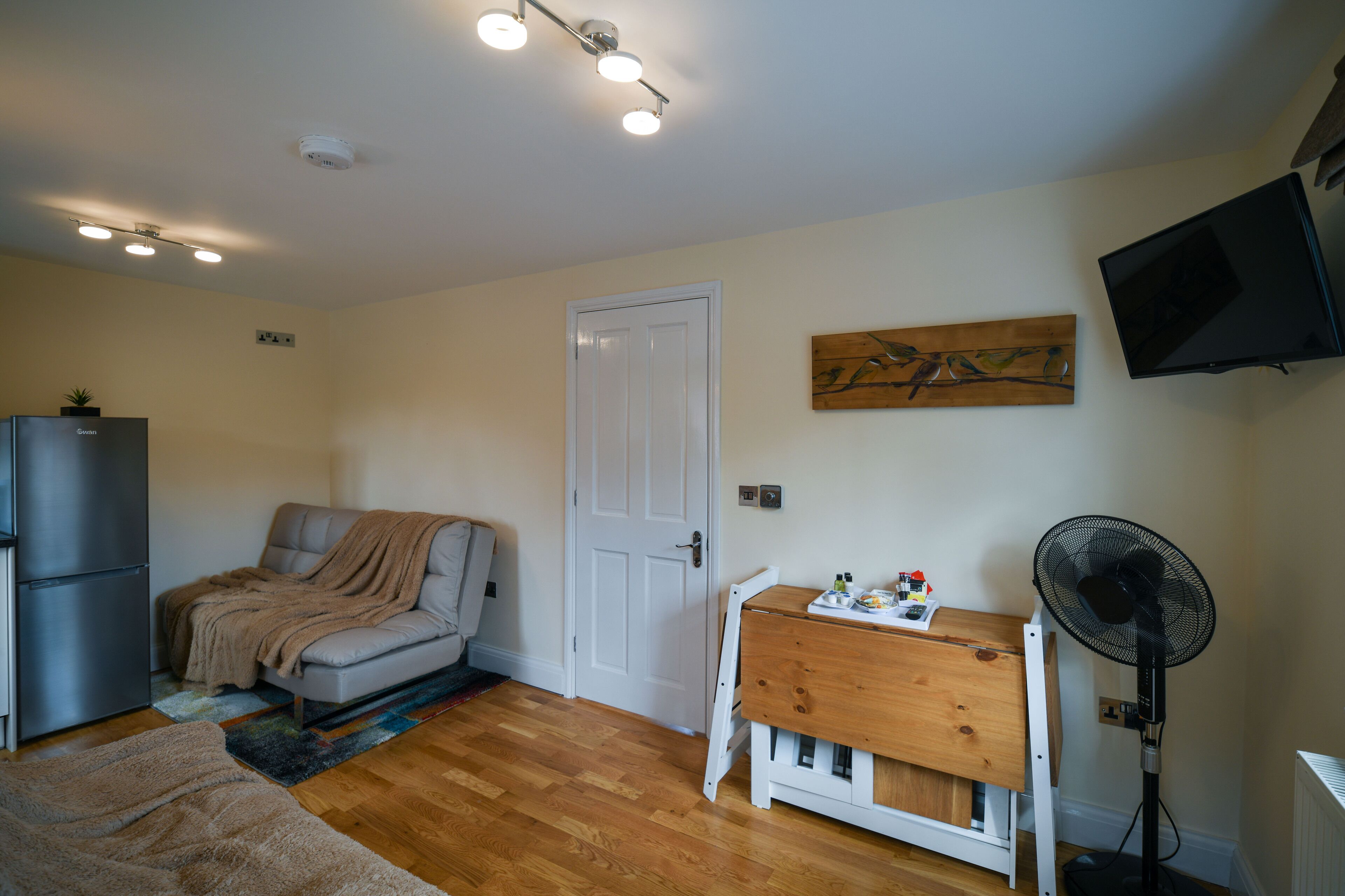 A compact living space with a sofa, a small wooden table, a fan, a TV, and a refrigerator.