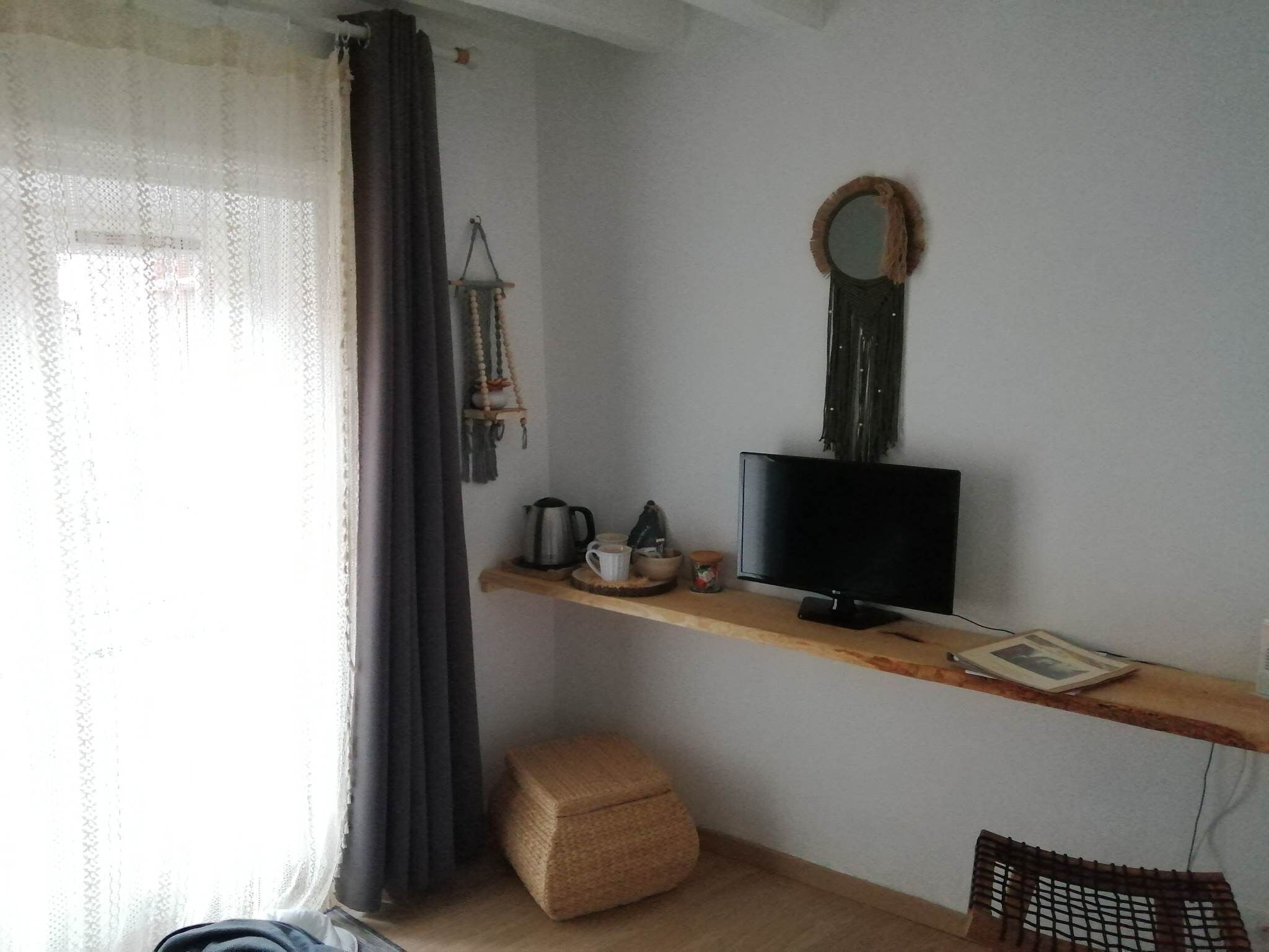 A room with a wooden shelf holding a television, a kettle, and a cup. There's a woven stool and a small basket on the floor.