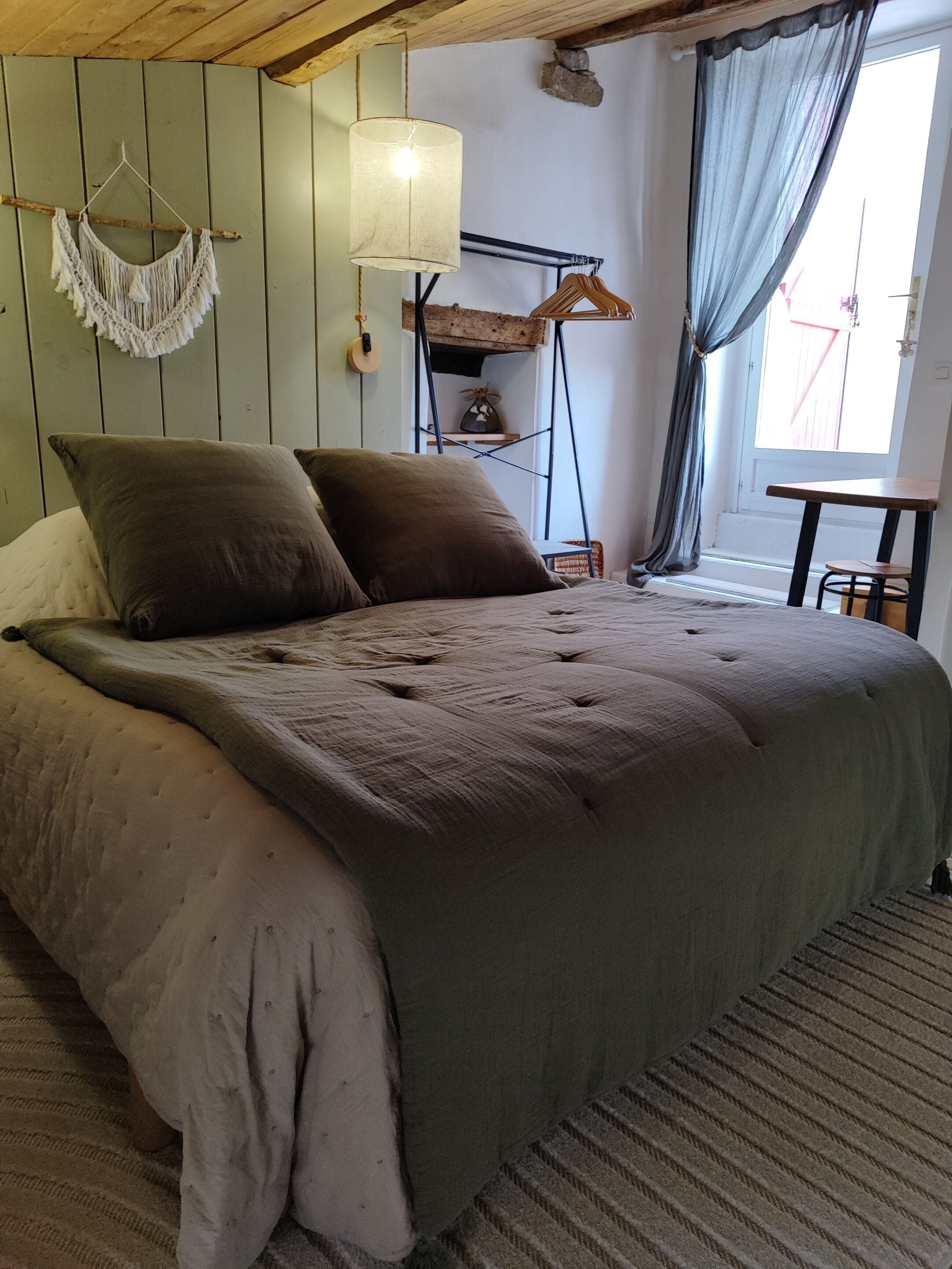 A bed with a grey quilt, a macrame wall hanging, a hanging light, and a rustic wooden shelf.
