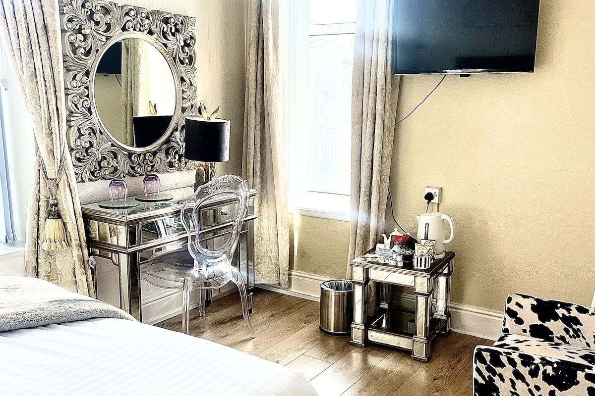A hotel room with a bed, a desk with a mirror, a television mounted on the wall, a window with curtains, and a small table with a kettle.