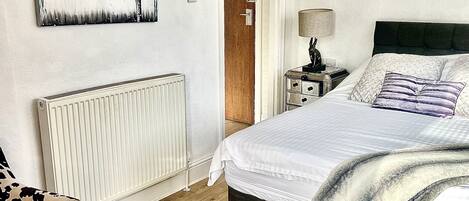 Quarto com cama, poltrona, radiador, porta e duas pinturas penduradas na parede.