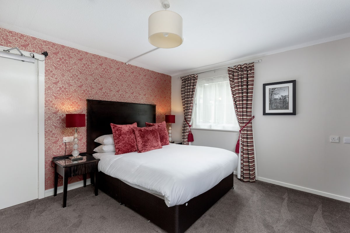 A bedroom with a large bed, a nightstand, a lamp, a window with curtains, and a wall-mounted picture.