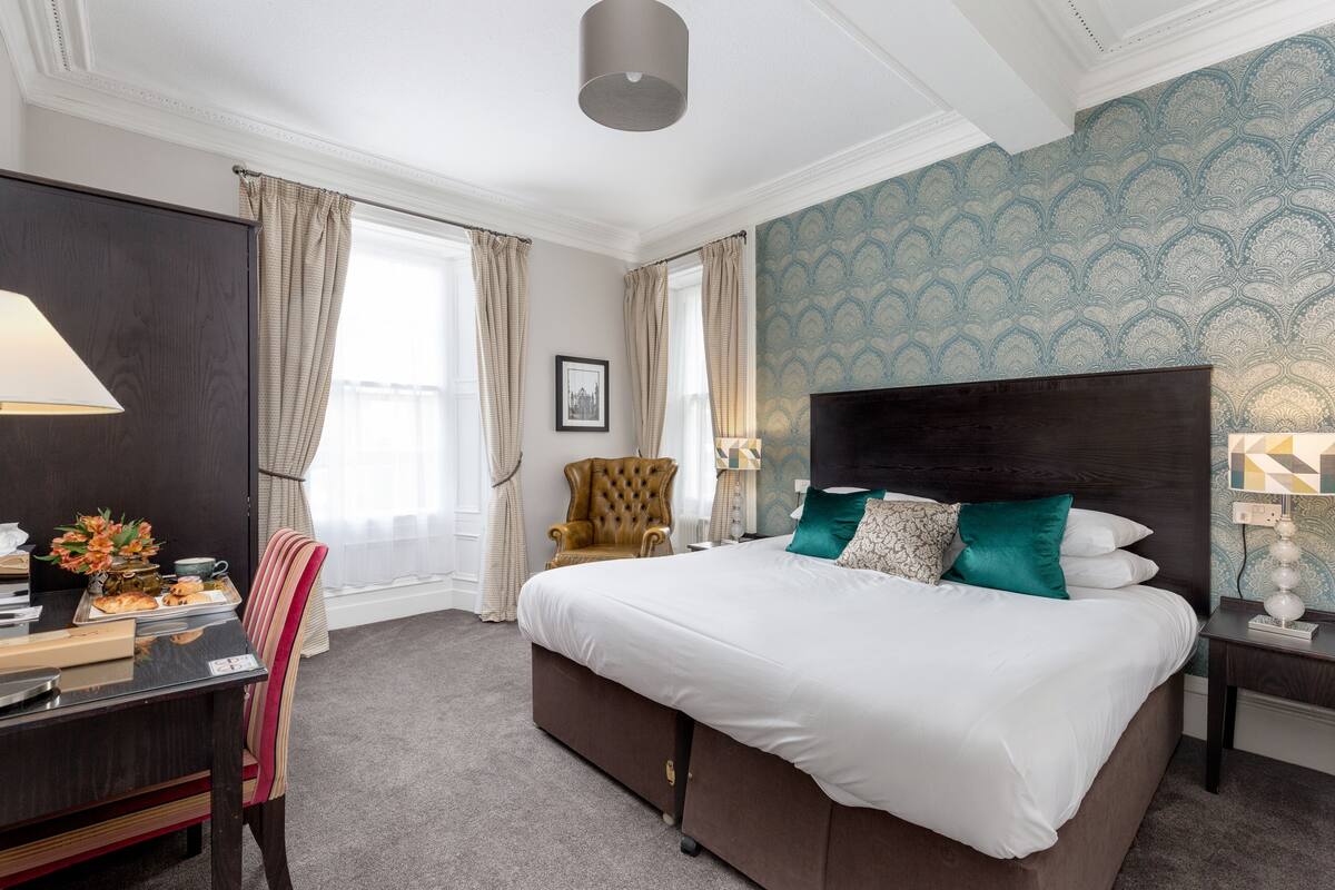 A hotel room with a large bed, a desk, a chair, and a window with curtains.