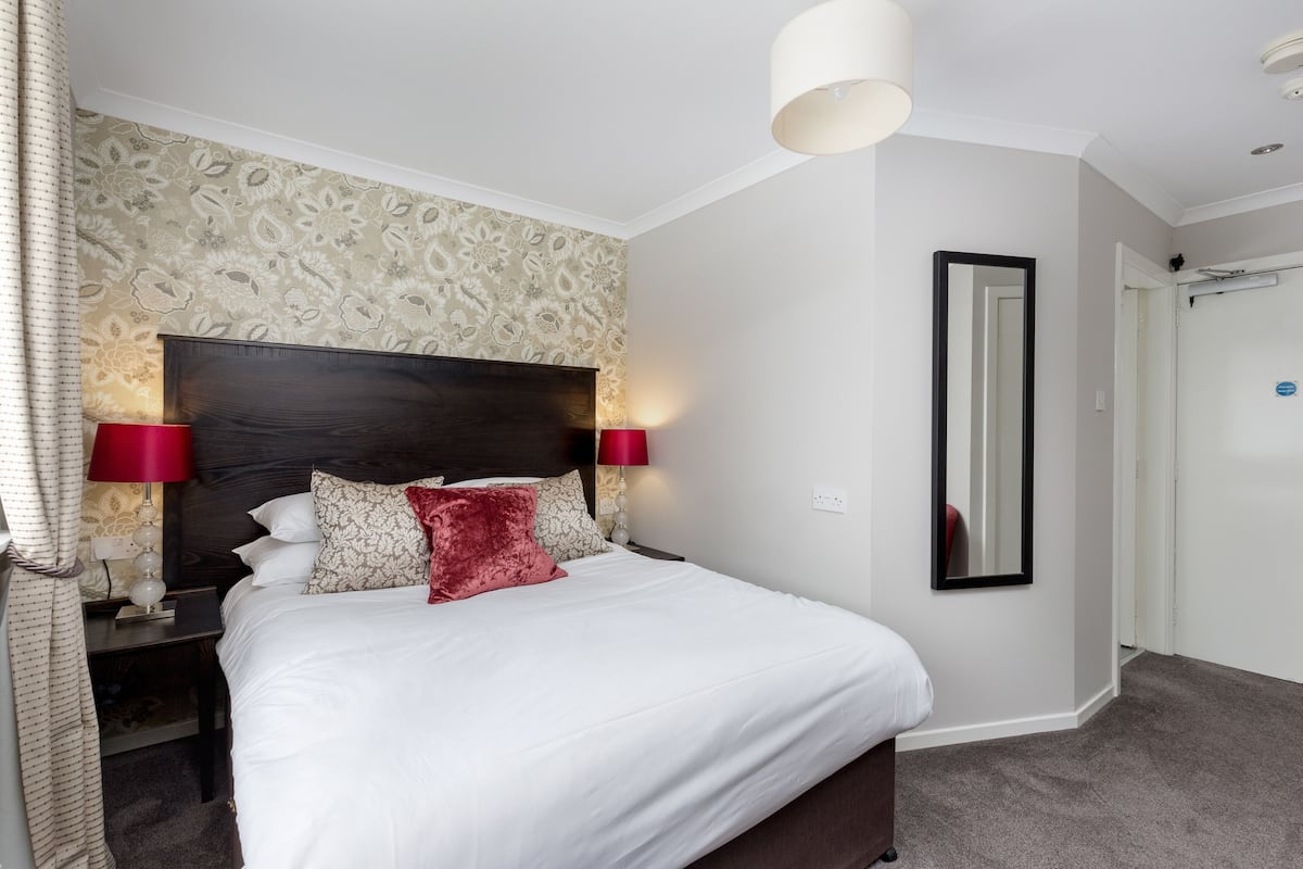 A neatly made bed with white linens and a red accent pillow, a dark wooden headboard, and a patterned wallpaper.