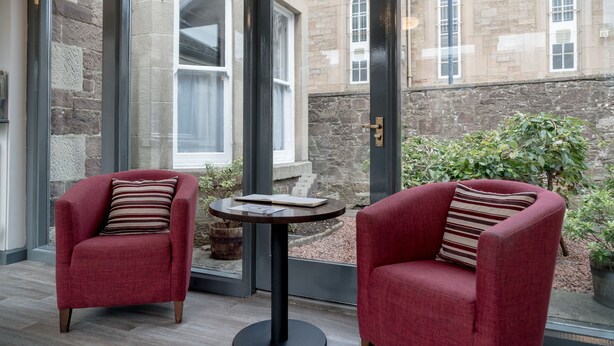 Two red armchairs with striped pillows, a small round table, and a view of a stone building with arched windows through a glass door. - The Kirkstyle Hotel (Biggar)