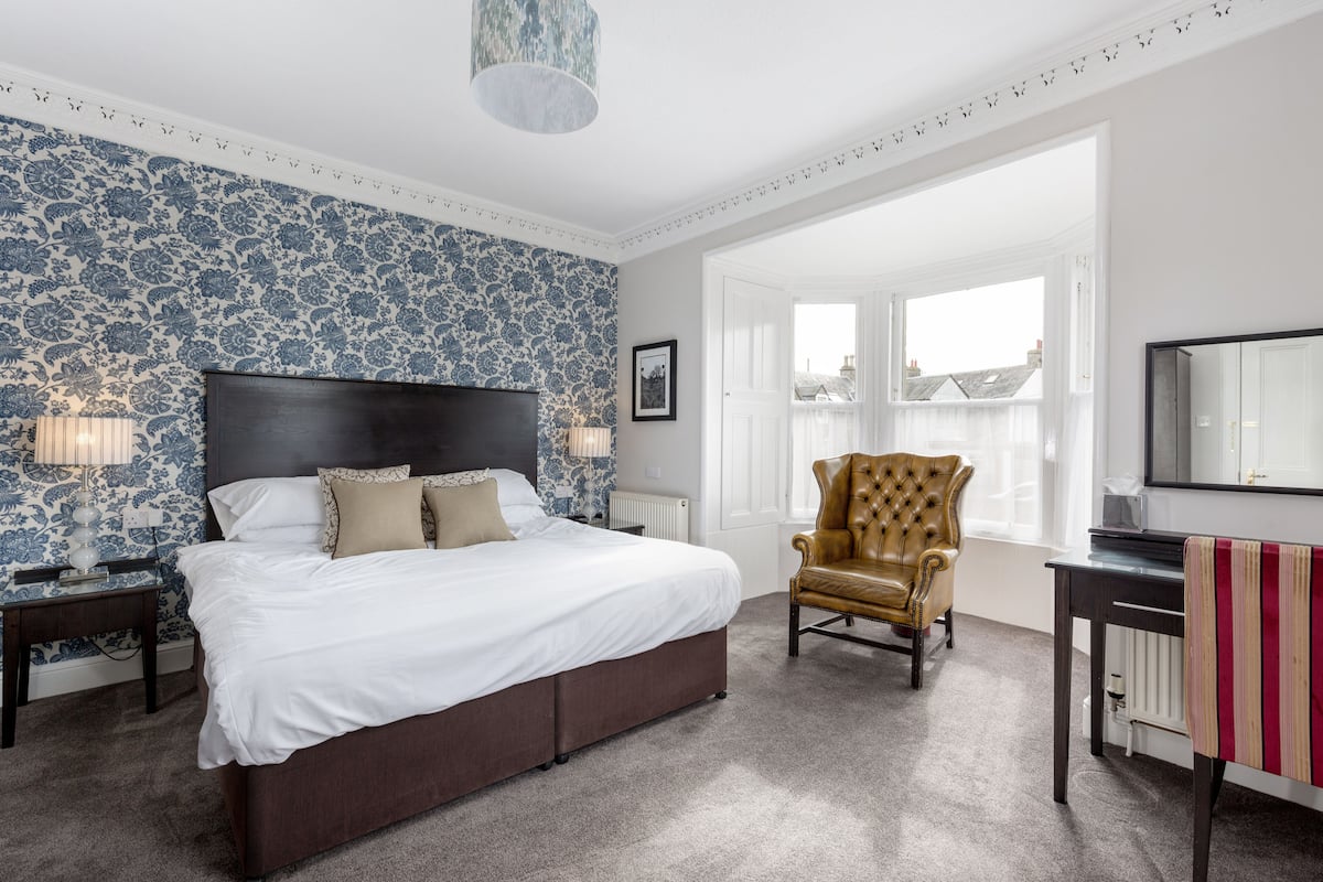 A bedroom with a large bed, a leather armchair, a desk, and a view of the city through large windows.