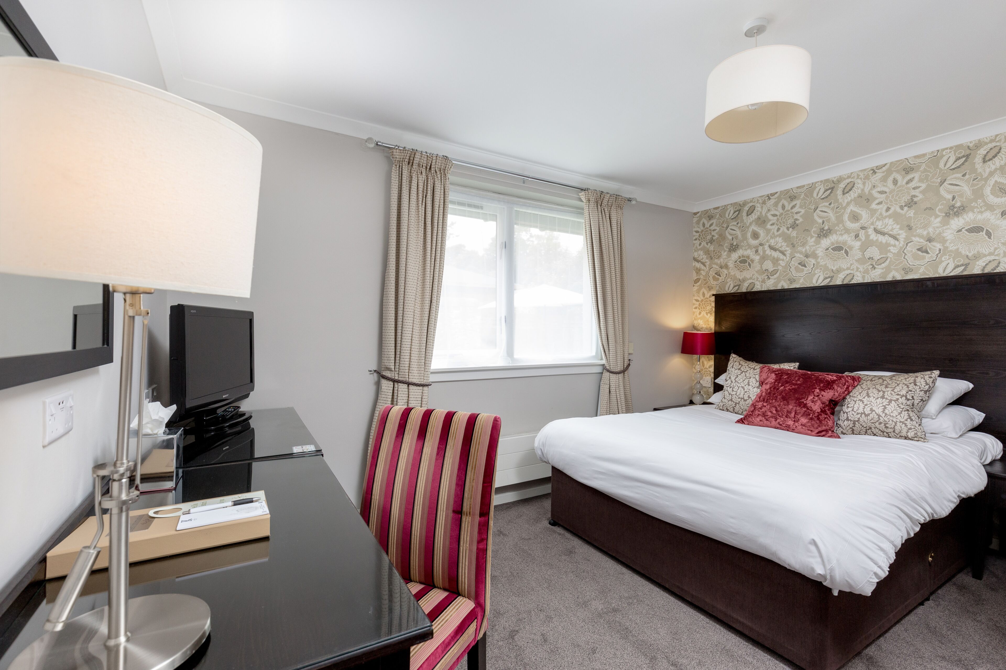 A hotel room with a bed, a desk with a television, a chair, and a window with curtains.