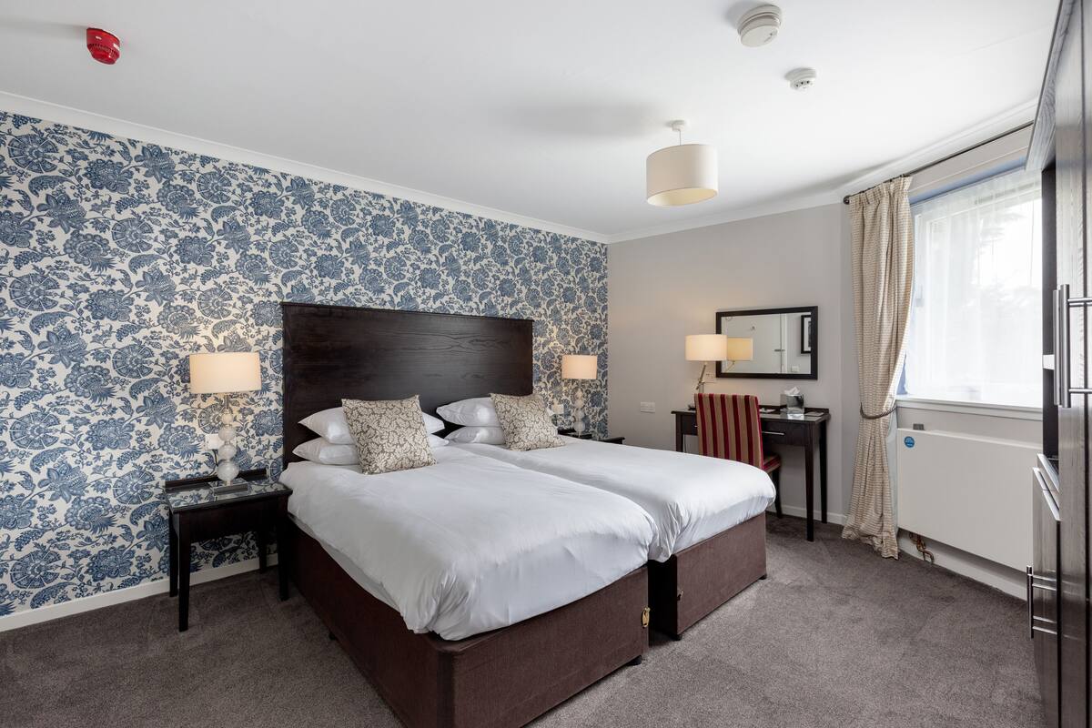 A double bed with a dark headboard, two bedside tables with lamps, a small desk with a chair, a radiator, and a window with curtains.