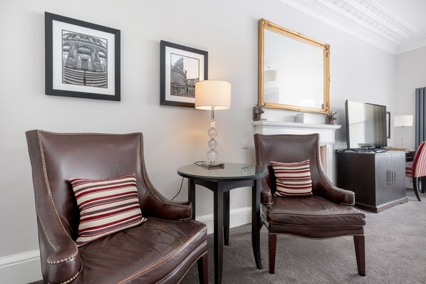 A room with two leather armchairs, a round table with a lamp, framed pictures on the wall, a large mirror, and a fireplace. - The Kirkstyle Hotel (Biggar)