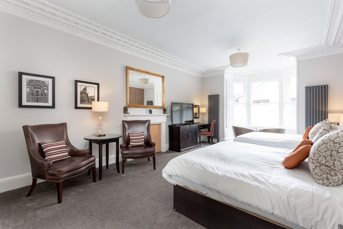 A hotel room with a large bed, two armchairs, a desk, a television, and framed pictures on the wall.