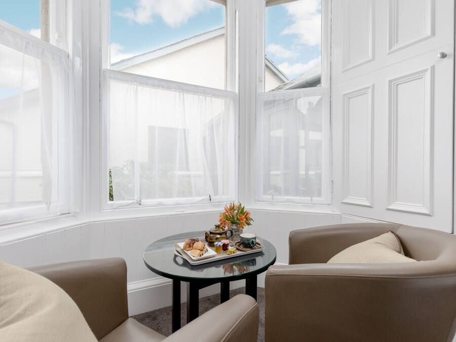 Una habitación con una mesa redonda dispuesta para el desayuno, dos sillones y ventanas amplias con contraventanas blancas.