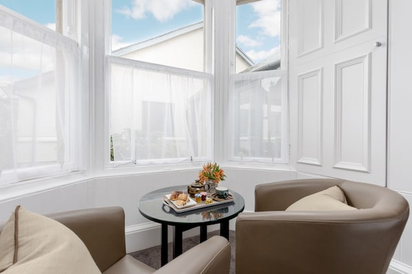 A room with a round table set for breakfast, two armchairs, and large windows with white shutters.