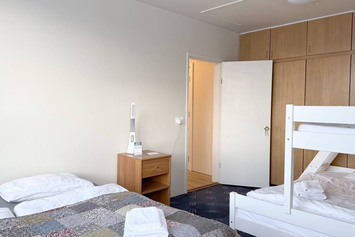 Ein Hotelzimmer mit Etagenbett, Nachttisch mit Lampe, kleinem Holfnachtkästchen und einer Tür zu einem weiteren Raum.