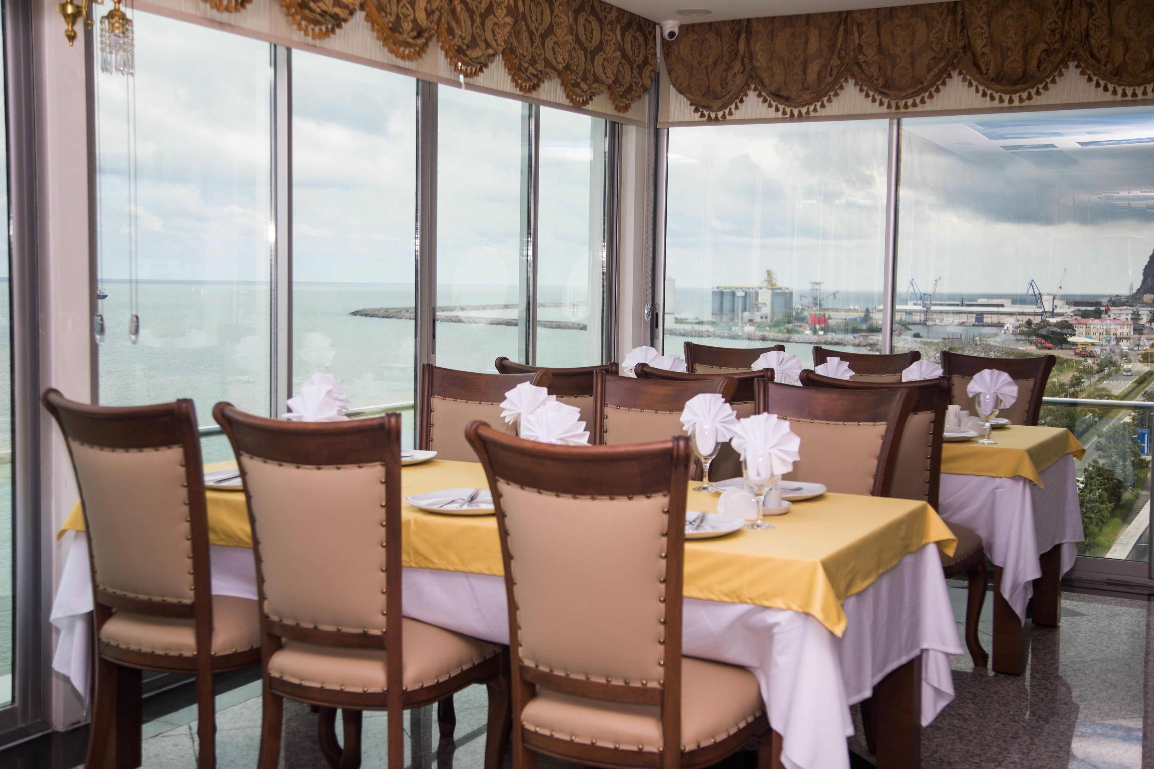 A dining area with tables set for service, large windows offering a view of the sea and cityscape.