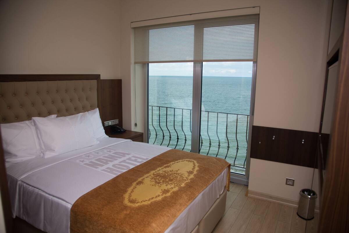 A hotel room with a large bed, a view of the sea through a balcony, and a window with blinds.