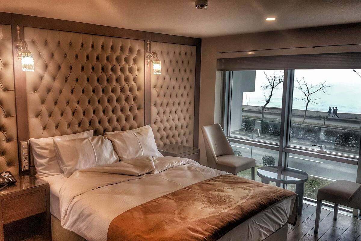 A modern hotel room with a tufted headboard, a large bed, a desk with a phone, a chair, and a view of the city through large windows.