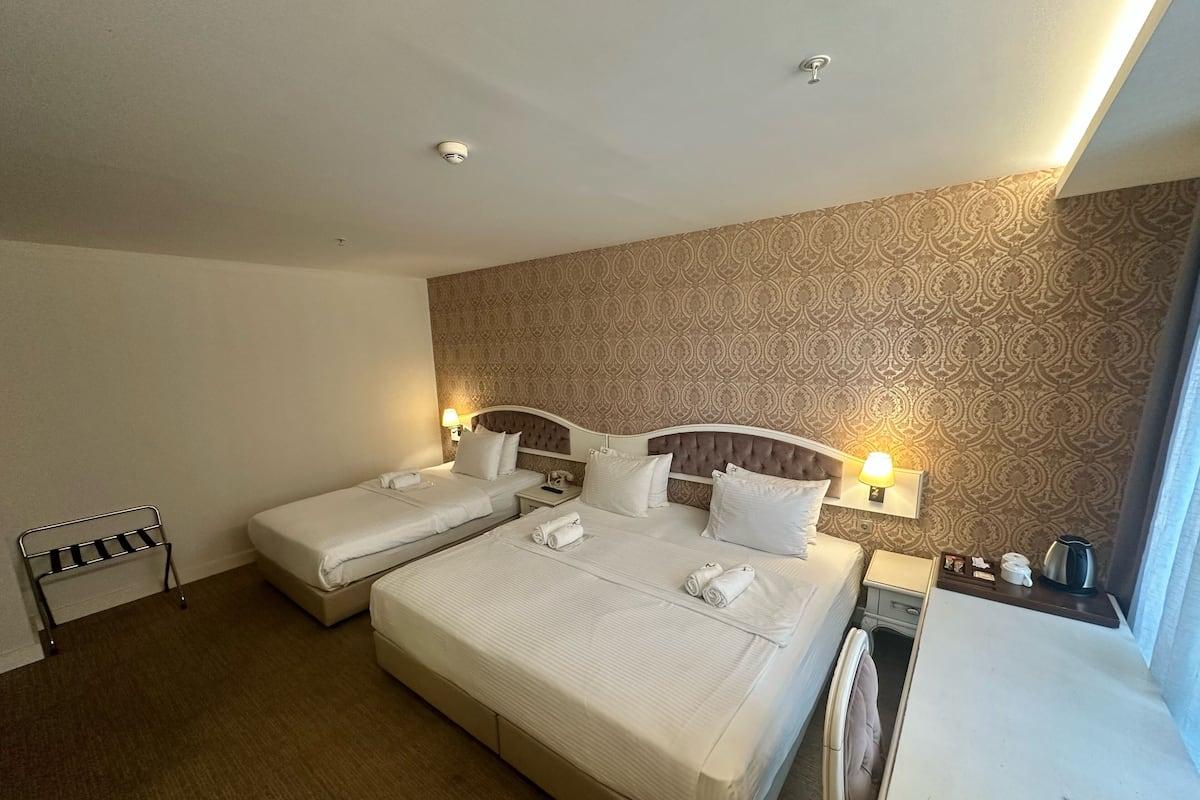 A hotel room with two beds, a bedside table with a lamp, a small table with a kettle, and a patterned wall.