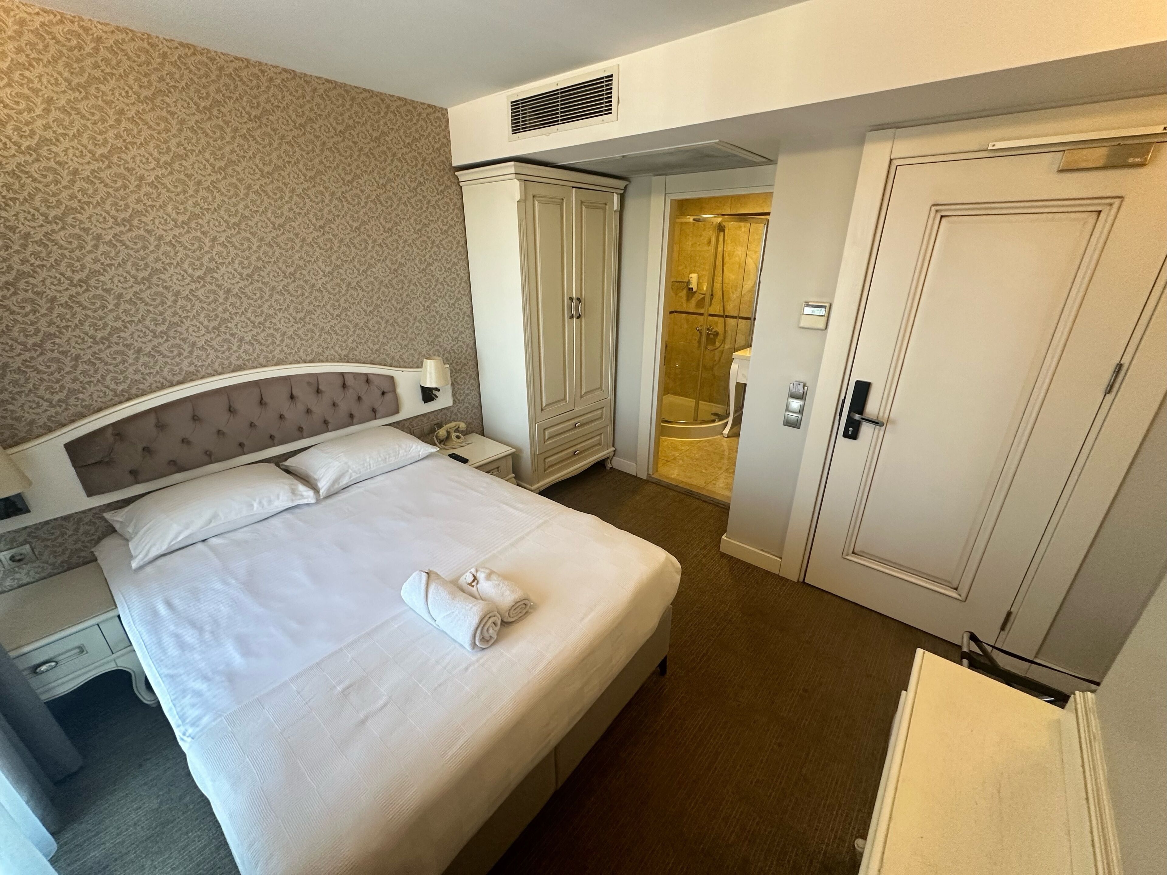 A hotel room with a bed, a wardrobe, a bathroom with a shower, and a bedside table with a lamp.
