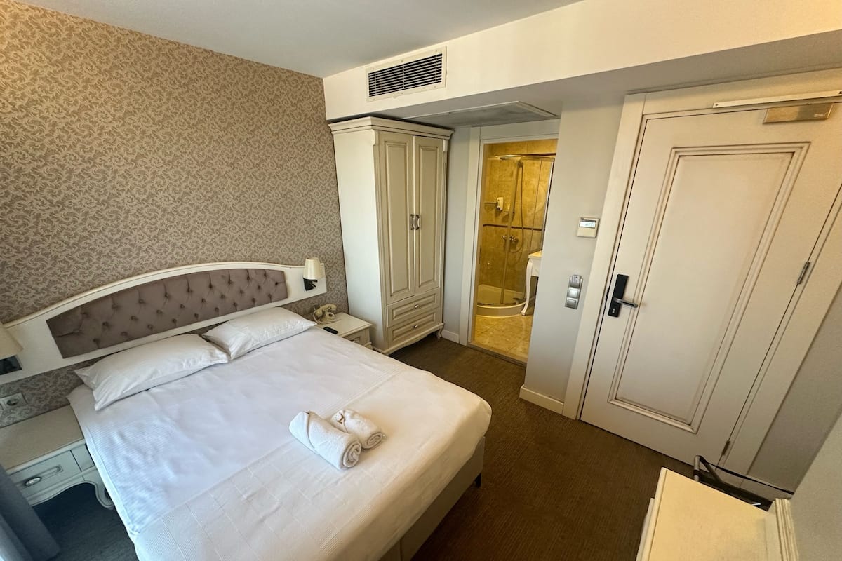 A hotel room with a bed, a wardrobe, a bathroom with a shower, and a bedside table with a lamp.