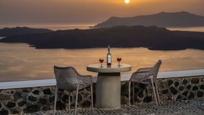 A sunset view from a stone-walled terrace with two wicker chairs and a table, featuring a bottle and two glasses of red wine. - Villa Thiranthemis - Adults Only (Santorini)
