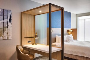 Habitación de hotel con escritorio, silla, cama con ropa blanca, mesita de noche y lámpara fijada en la pared.