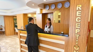 A reception desk with a wooden finish and a sign reading "RECEPTION". There are three clocks above the desk, and a menu board to the right. - Sonerbey Otel (Ordu)