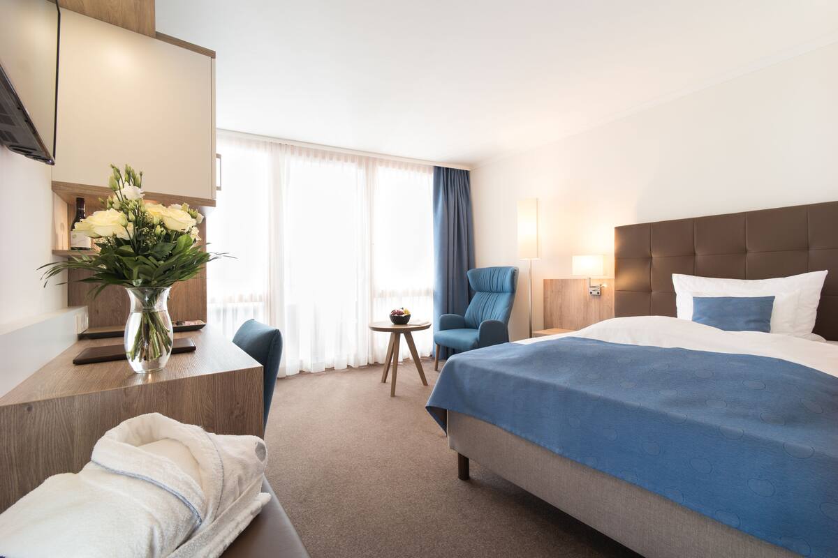 A hotel room with a large bed, a desk with a vase of flowers, a chair, and a small table with a bowl of fruit.