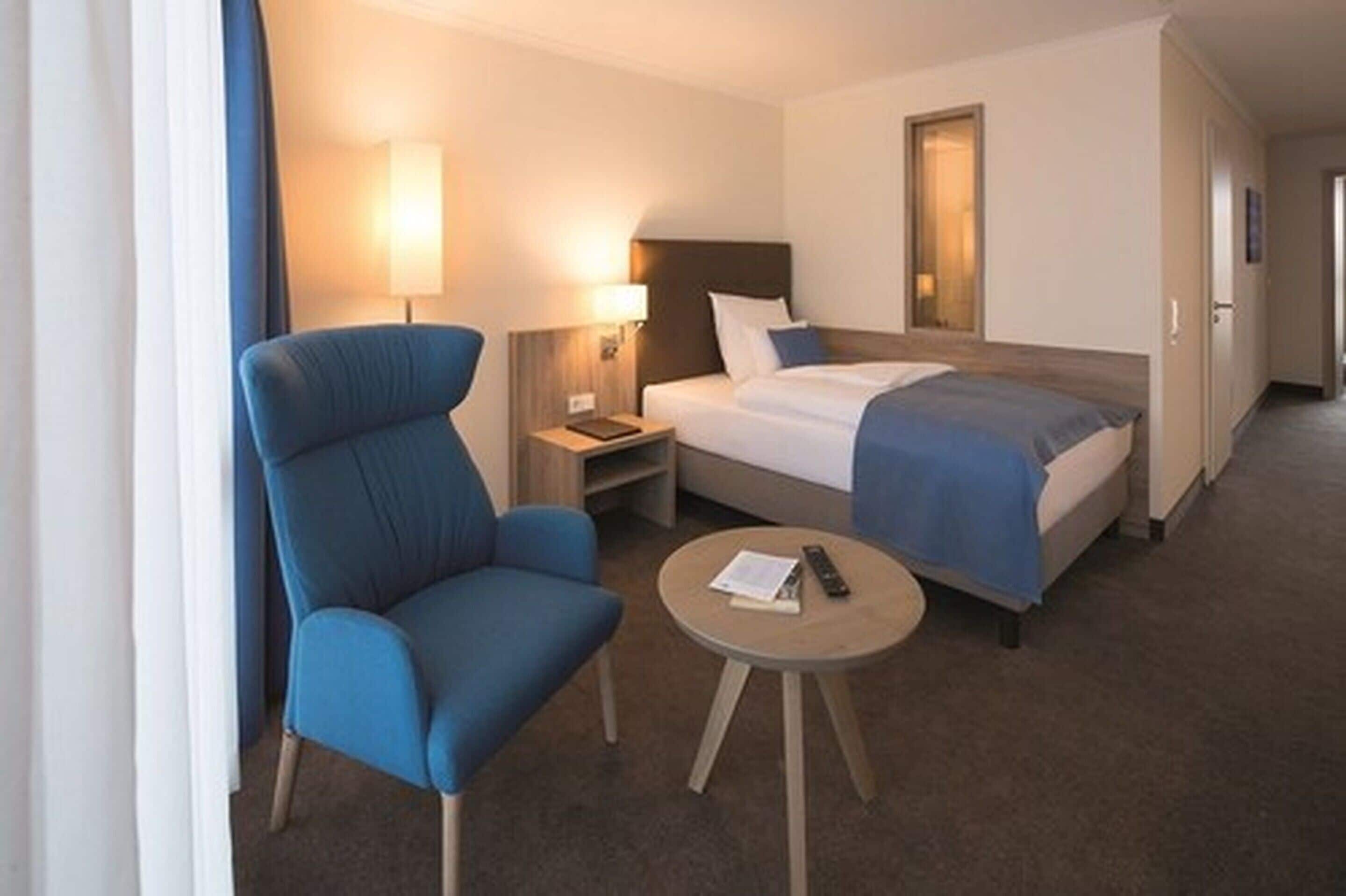 A hotel room with a large bed, a blue armchair, a bedside table with a lamp, and a small round table with a remote control and a book.