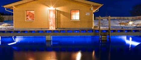 A wooden cabin with a pool in front, illuminated by lights at night.