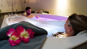 A person relaxing in a bathtub with a towel and flowers, another person in the background, and a chandelier. - Siebenquell GesundZeitResort (Weissenstadt)