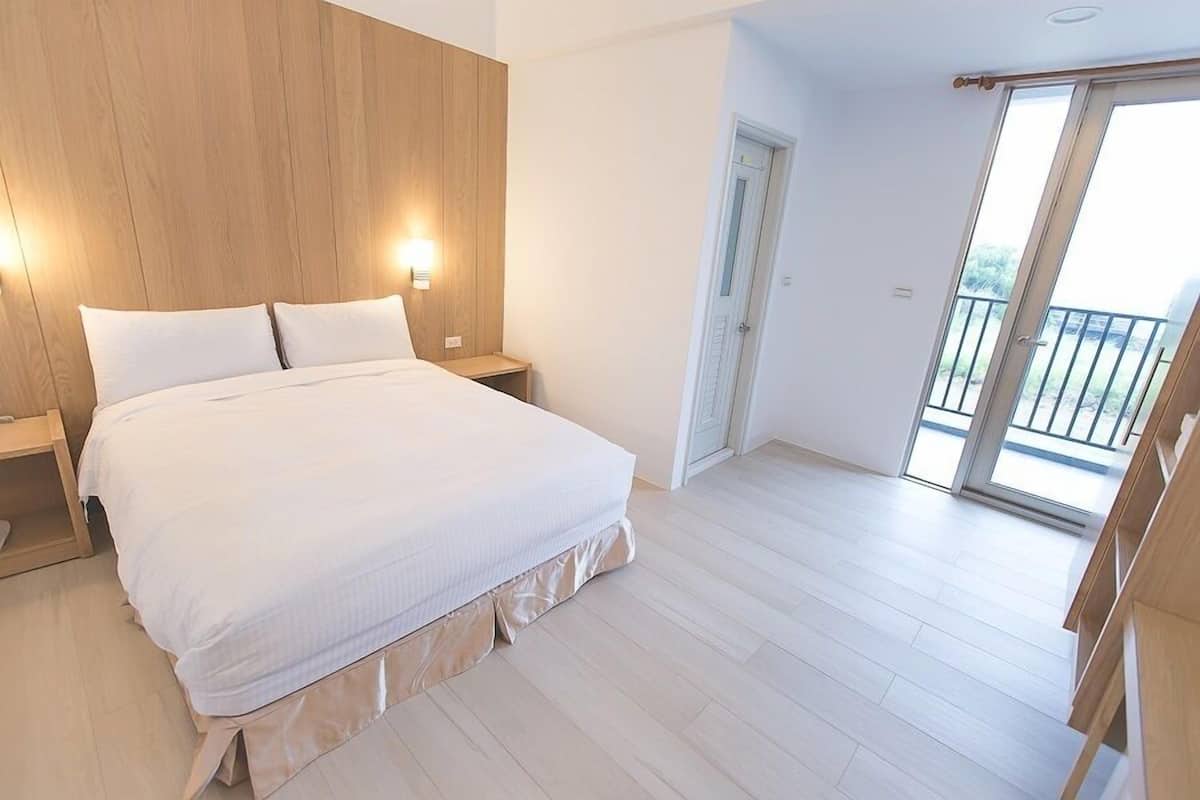 A bedroom with a large bed, wooden headboard, bedside lamps, a sliding glass door to a balcony, and a wooden floor.
