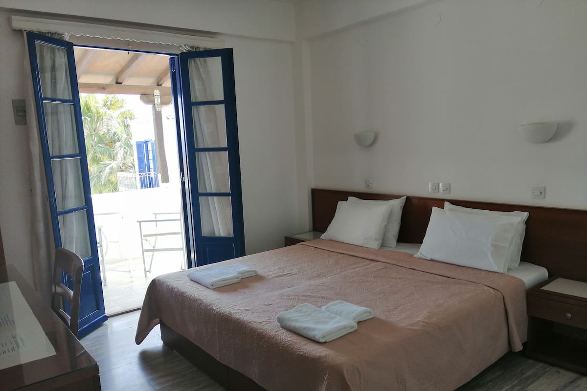 A hotel room with a bed, a nightstand, a desk, and a chair. There is a window with curtains, and a door leading to an outdoor area.