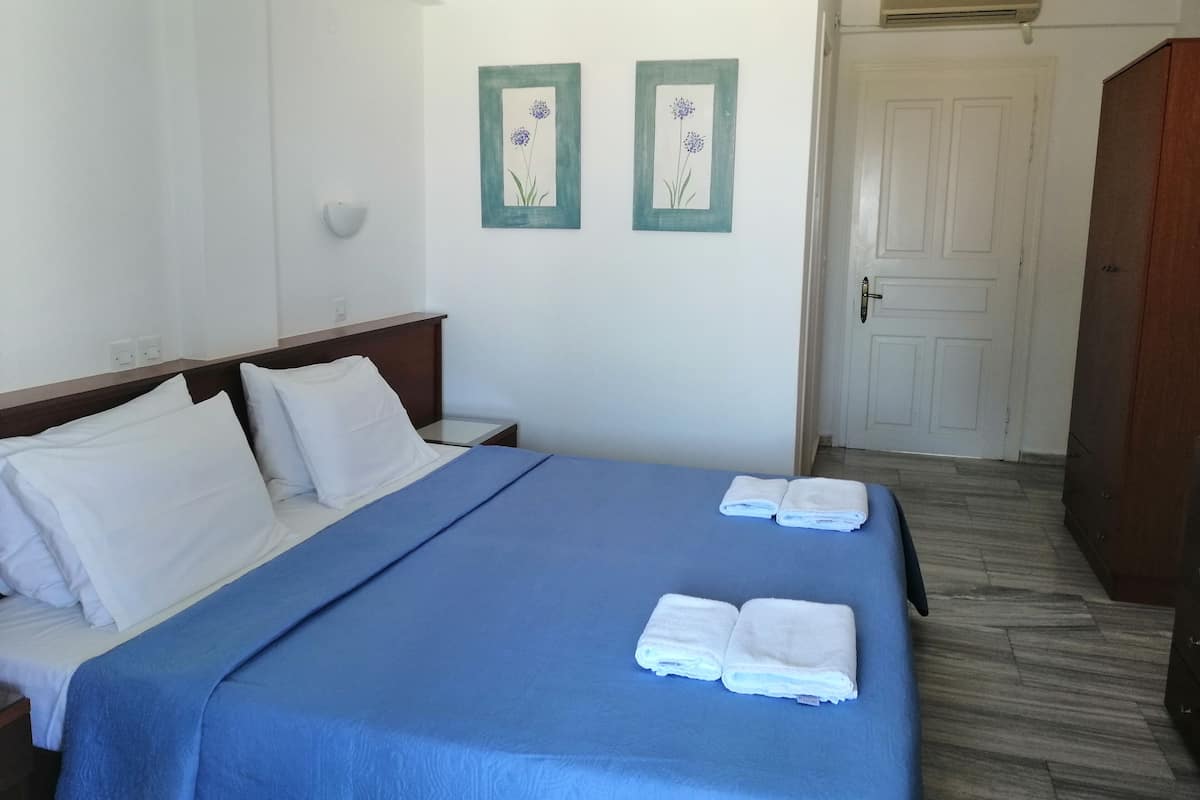 A hotel room with a bed, white pillows, a blue bedspread, two wall-mounted air conditioning units, and a door.