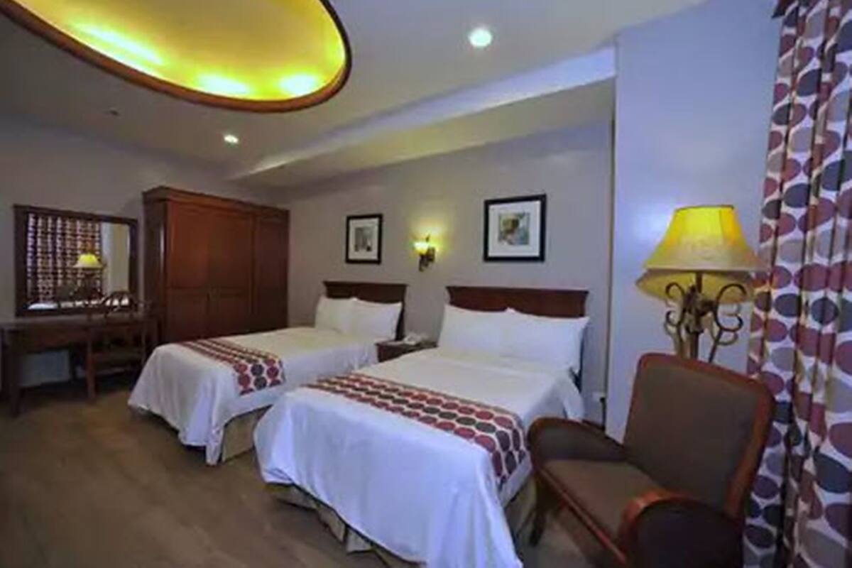 A hotel room with two beds, a desk, a chair, and a lamp.