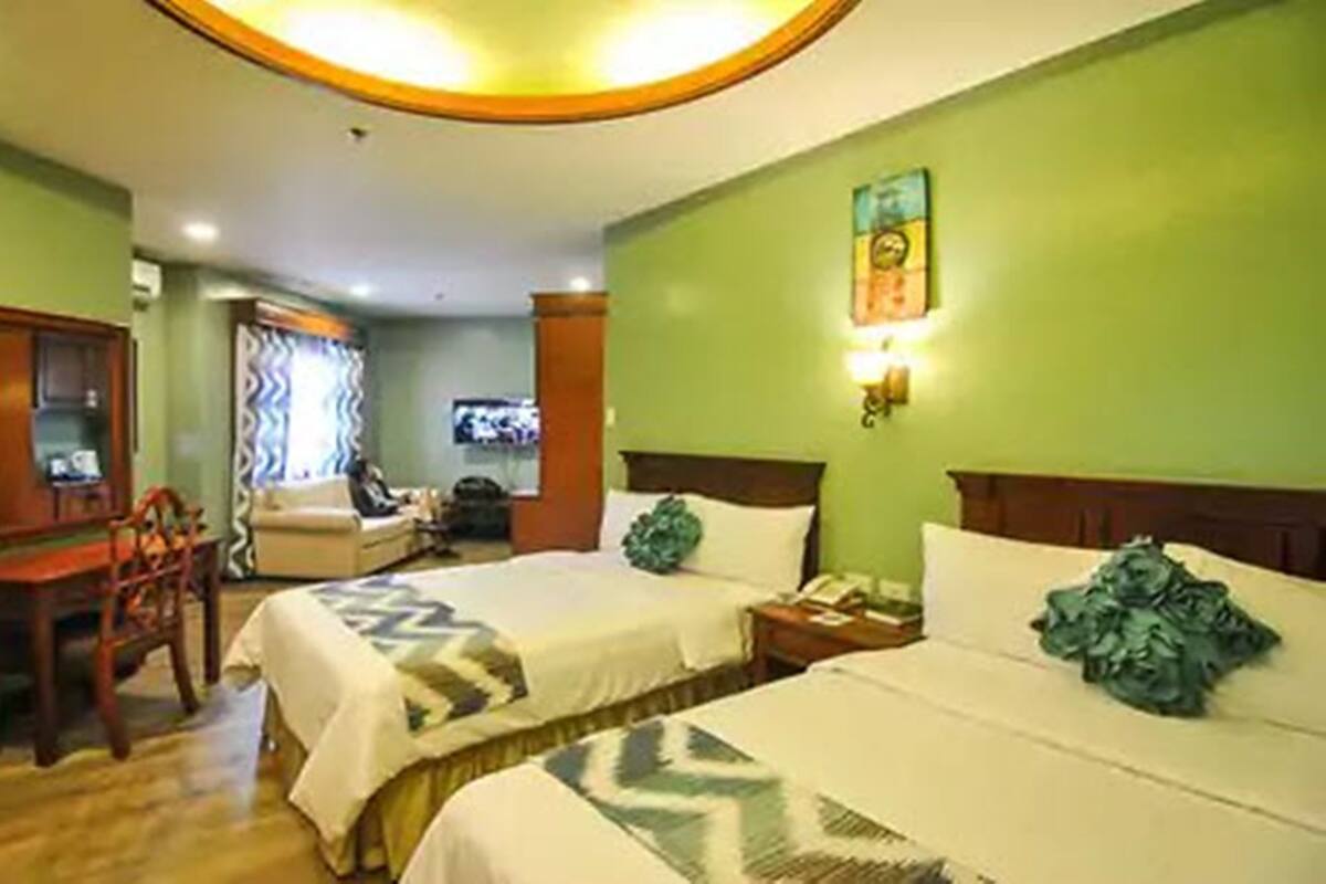 A hotel room with two beds, a desk, a chair, a television, and a wall-mounted light fixture.